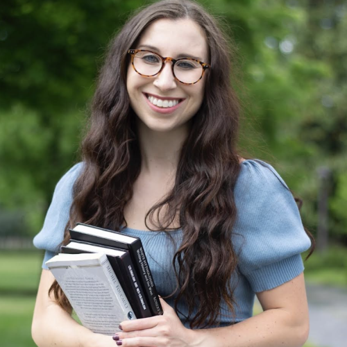 Courtney in a blue top, holding books and smiling at camera